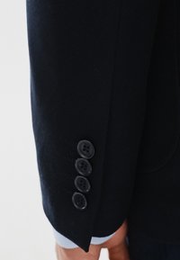 Marynarski żakiet z rękawem wykonanym z teksturowanej tkaniny, czterema czarnymi guzikami oraz kontrastowym jasnoniebieskim wykończeniem mankietu.