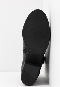 Semelle noire d'une chaussure avec un motif de grip texturé, présentée debout verticalement contre un fond blanc en coin.