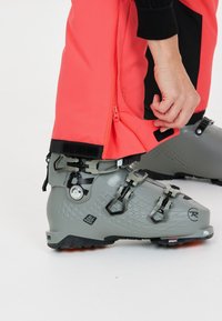 Grijze skiboot met een gestructureerd oppervlak, voorzien van zwarte hardware en verstelbare gespen, naast een helderroze broek met ritsen en klittenbandsluitingen.
