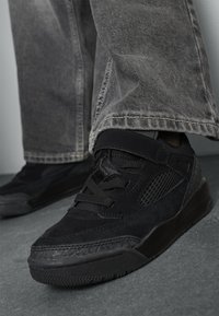Baskets noires avec un mélange de daim et de matériau en mailles, présentant un col rembourré, une fermeture à lacets et une semelle texturée. Portées avec un jean en denim gris.