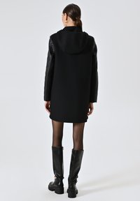 Cappotto nero con cappuccio e corpo texturizzato, con maniche in pelle. Indossato con collant neri trasparenti e stivali neri fino al ginocchio.