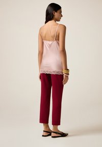 Rosa Satin-Cami mit Spitzenbesatz, kombiniert mit bordeauxfarbenen schmalen Hosen und schwarzen flachen Sandalen. Model zeigt Rückansicht. Schlichte Accessoires.