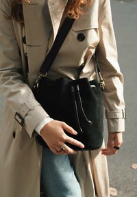 Schwarze Wildleder-Beuteltasche mit Kordelzugverschluss, ausgestattet mit goldfarbenen Beschlägen und einem schwarzen verstellbaren Riemen, gehalten von einer Person in einem beigen Trenchcoat.