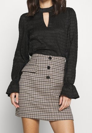 Vrouw draagt een zwarte gestructureerde blouse met lange mouwen, sleutelgatnek en een hoge taille pied-de-poule minirok met knopen.