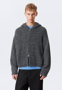 Joven vestido con una sudadera gris de felpa con cremallera sobre una camisa azul y pantalones negros, de pie contra un fondo blanco liso.
