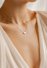 Silberne Halskette mit einem kleinen Herz-Anhänger, versehen mit einer zarten Kette. Die Trägerin hat einen nackten Hals und trägt ein leichtes, strukturiertes Oberteil.