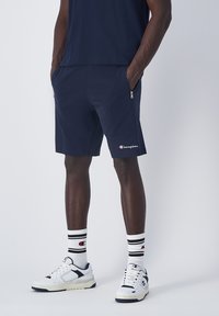 Pantalones cortos deportivos de color navy con bolsillos laterales con cremallera, que presentan un pequeño logo blanco. Combinados con zapatillas blancas y calcetines a rayas.