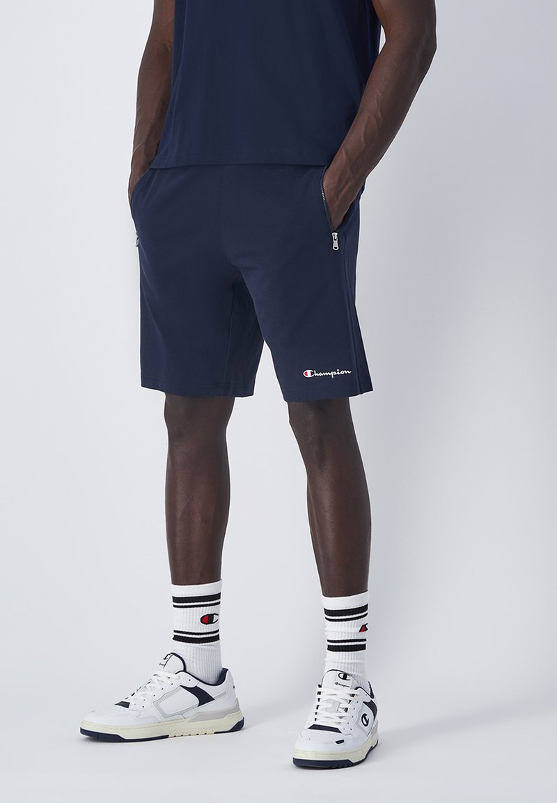 Pantalones cortos deportivos de color navy con bolsillos laterales con cremallera, que presentan un pequeño logo blanco. Combinados con zapatillas blancas y calcetines a rayas.