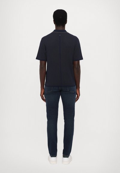 Marine stripete kortermet polo med krage, kombinert med mørke denimjeans og hvite sneakers, sett fra baksiden.