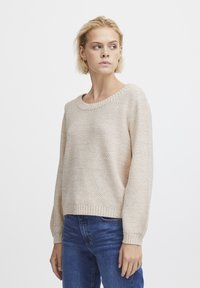 Hellbeige gestrickter Pullover mit lockerer Passform, Rundhalsausschnitt und gerippten Bündchen, kombiniert mit blauen Jeans. Subtiles strukturiertes Muster zu erkennen.