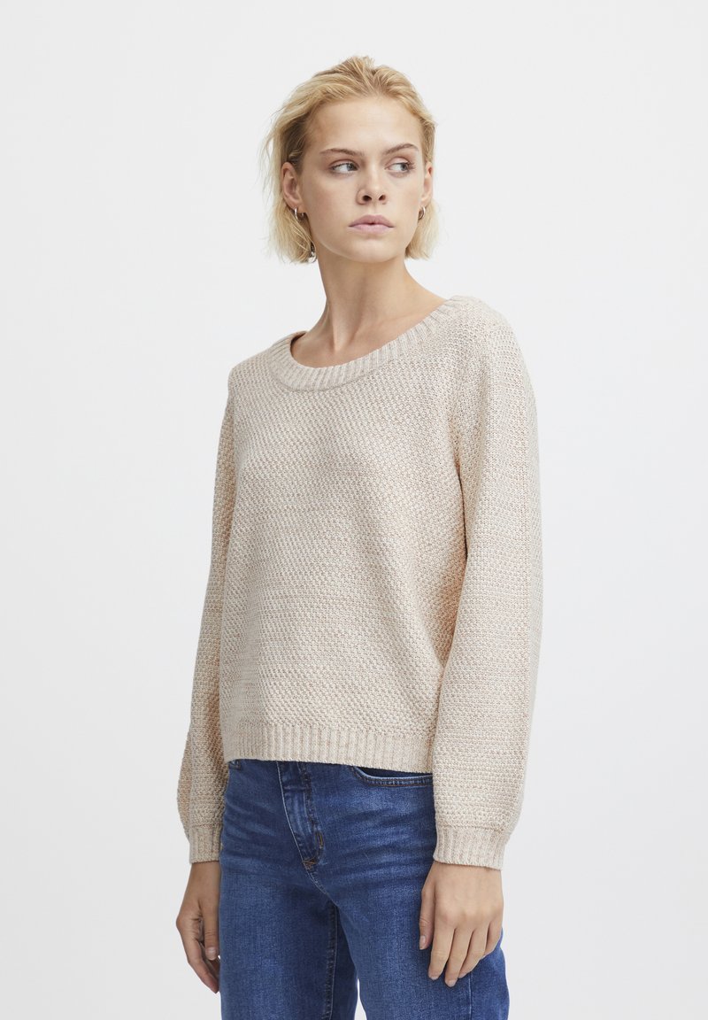 Hellbeige gestrickter Pullover mit lockerer Passform, Rundhalsausschnitt und gerippten Bündchen, kombiniert mit blauen Jeans. Subtiles strukturiertes Muster zu erkennen.