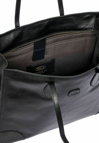 Zwarte leren tote bag met platte handvaten, open bovenkant en een grijze binnenkant. Heeft een ritsvak en een merkembleem aan de binnenkant.