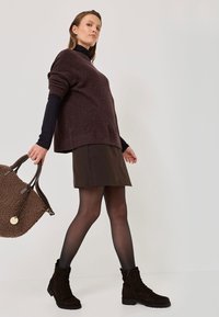 Brauner Oversized-Pullover, schwarzer Rollkragenpullover, brauner Minirock, schwarze Strumpfhose und dunkelbraune Schnürstiefeletten; trägt eine strukturierten braune Handtasche.