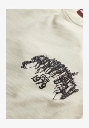 T-shirt di cotone beige chiaro con testo grafico nero che recita "Supertramp Tour 1979" con un design stilizzato.