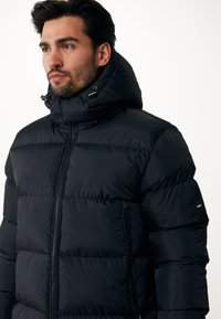 Chaqueta acolchada negra con cuello alto y cierre de cremallera. Presenta un diseño acolchado segmentado, capucha con cordón y bolsillos laterales con cremalleras.