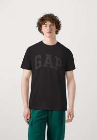 T-shirt noir à manches courtes avec un grand logo "GAP" gris foncé sur le devant ; tissu doux avec un design classique à col rond.
