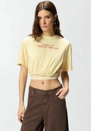 Giovane donna che indossa una maglietta corta gialla con scritta e pantaloni marroni a vita alta, in piedi con una mano in tasca su sfondo bianco.