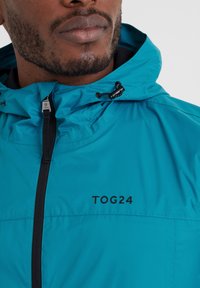 Teal waterproof jacket with a hood, featuring a black front zipper and "TOG24" logo. Smooth texture and adjustable drawstring at the neck.