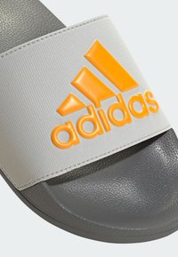 Gray slide sandal featuring a textured strap with an orange raised "adidas" logo. Soft rubber sole with a smooth finish.