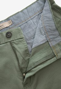 Olivgrüne gerade geschnittene Hose mit Reißverschluss, Knopfverschluss und einem kontrastierenden gestreiften Innenfutter mit aufgenähtem Etikett.