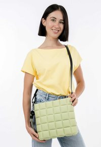 Borsa a mano verde di forma quadrata con un motivo trapuntato testurizzato, tracolla nera, tenuta da un modello che indossa una camicia gialla e jeans blu.