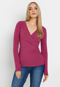 Mauve Langarmoberteil mit tiefem V-Ausschnitt und Wickeldesign, aus glattem Stoff gefertigt und figurbetont geschnitten, kombiniert mit blauen Jeans.