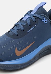 Niebieski but biegowy Nike z teksturowaną cholewką, z brązowym logo Swoosh, wzmocnioną czubkiem i sznurowanym zapięciem. Podeszwa z bieżnikiem do trudnych warunków pogodowych.