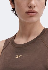 Gros plan d’une personne portant un haut marron sans manches avec un logo Reebok doré sur la poitrine, montrant une partie du cou et du bas du visage.