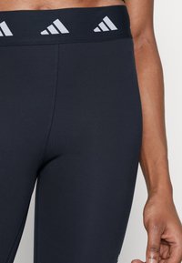 Leggings sportivi neri con vita alta, caratterizzati da accenti del logo Adidas grigio chiaro e una texture di tessuto liscia e elastica.