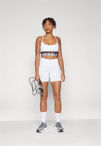 Baltas sportinis liemenukas su juodu "NEBBIA" logo, derinamas su baltais aukštu juosmeniu šortais. Modelis laiko šokinėjimo virvę ir dėvi pilkas sportbačius.