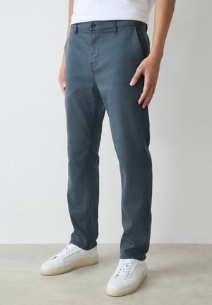 Person iført mørkeblå lige benbukser og hvide sneakers, der står foran en enkel lys baggrund.