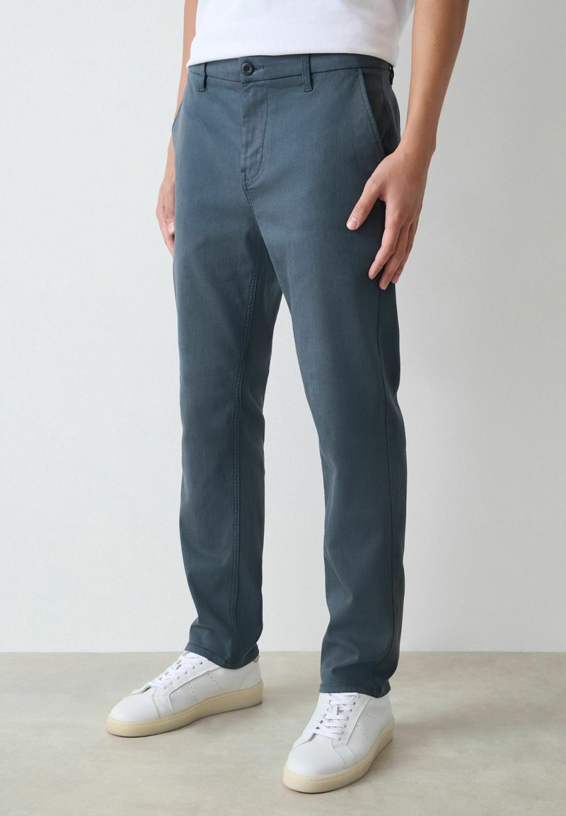 Sötétkék, egyenes szárú nadrágot viselő személy fehér sportcipővel, egy sima, világos háttér előtt áll.