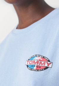Camiseta de algodón azul claro con un logo circular en negro, rojo y azul que dice "TOMMY JEANS" y un diseño de palmera. Mangas cortas.