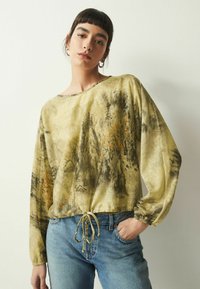 Langärmliges Oberteil aus leichtem, strukturiertem Stoff. Mit einem abstrakten Muster in Grün und Braun, Kordelzugsaum und lockerer Passform. Kombiniert mit blauen Jeans.
