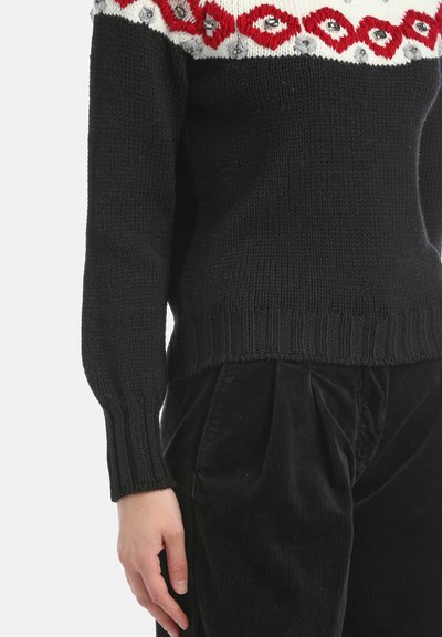Crni pleteni džemper s crveno-bijelim uzorkom na gornjem dijelu, uparen s crnim kordiranim hlačama visokog struka, prikazano od sredine trupa do ruke.