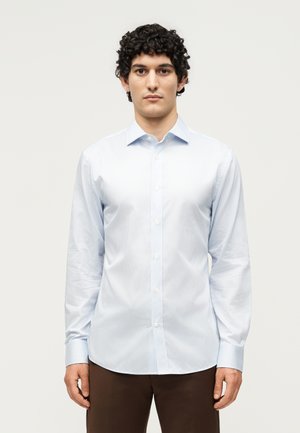 Mężczyzna z kręconymi włosami, ubrany w jasnoniebieską dopasowaną koszulę i brązowe spodnie, stoi na tle jednolitej białej ściany.