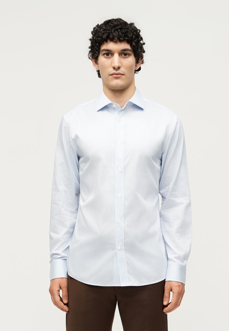 Άνδρας με σγουρά μαλλιά, που φοράει ένα ανοιχτό μπλε πουκάμισο στενής γραμμής και καφέ παντελόνι, στέκεται μπροστά σε ένα απλό λευκό φόντο.