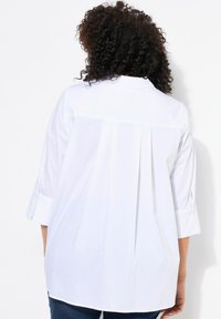 Camicia bianca con bottoni, dotata di una piega sul retro, con maniche arrotolate e una texture liscia. Progettata per una vestibilità rilassata.