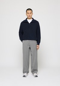 Pulover tricotat bleumarin cu guler și fermoar pe jumătate, asociat cu pantaloni largi gri deschis și adidași albi cu accente.