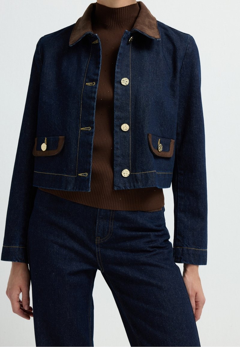 Personne portant une veste en jean bleu foncé et un jean assorti, avec un col marron et une bordure de poche, sur un pull col roulé côtelé marron.