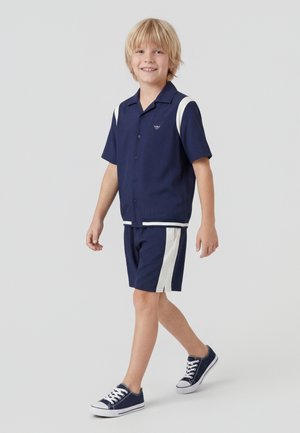 Αγόρι που περπατά, φορά μπλε σκούρο κοντομάνικο πουκάμισο και αντίστοιχα σορτς με λευκές ρίγες, σε συνδυασμό με μπλε σκούρα αθλητικά παπούτσια.