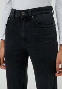 Jeans de mezclilla negra con cintura alta, bolsillos deshilachados y cierre de botón metálico; tejido texturizado; estilo ajustado con sutiles detalles de costura.