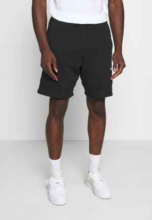 Homme portant un short noir à cordon, des chaussettes blanches montantes et des baskets blanches, debout devant un fond uni gris clair.
