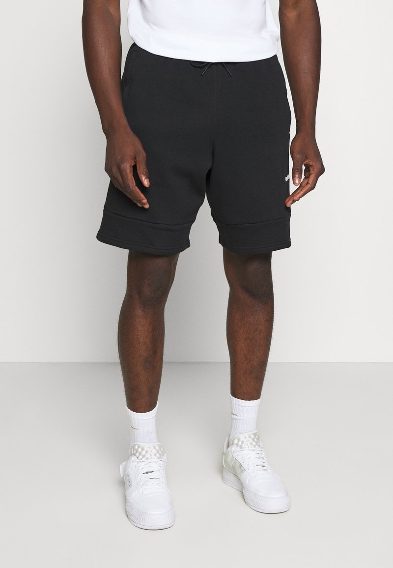 Homme portant un short noir à cordon, des chaussettes blanches montantes et des baskets blanches, debout devant un fond uni gris clair.