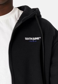 Sudadera con cremallera negra hecha de material suave, con un bolsillo frontal y un logo bordado en blanco y azul. Lleva una camiseta blanca debajo.