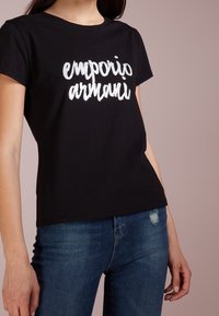 Svart bomullströja med "emporio armani" i vit brodyr, med klassisk rund hals och avslappnad passform. Matchas med blå denim.