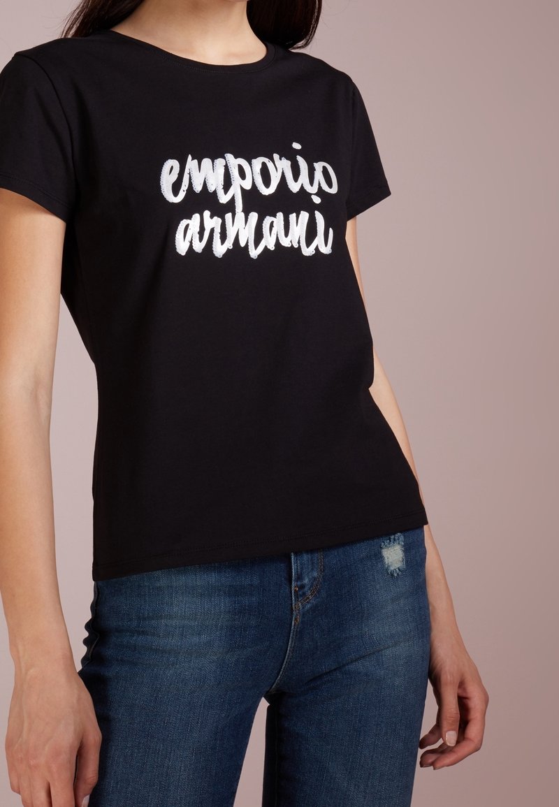 Svart bomullströja med "emporio armani" i vit brodyr, med klassisk rund hals och avslappnad passform. Matchas med blå denim.