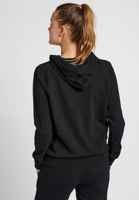 Czarna bluza z kapturem wykonana z miękkiego materiału, o luźnym kroju, z regulowanym kapturem i ściągaczami na rękawach, noszona z pasującymi spodniami.