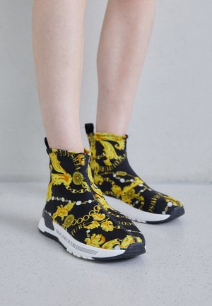 Jambes portant des baskets slip-on noires avec des motifs baroques jaunes ornés et des semelles blanches, debout sur un sol gris clair tacheté.