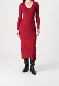 Robe rouge à manches longues en côtes, avec un décolleté arrondi, une silhouette ajustée et une longueur jusqu'aux chevilles, associée à des bottes noires montant jusqu'aux genoux.